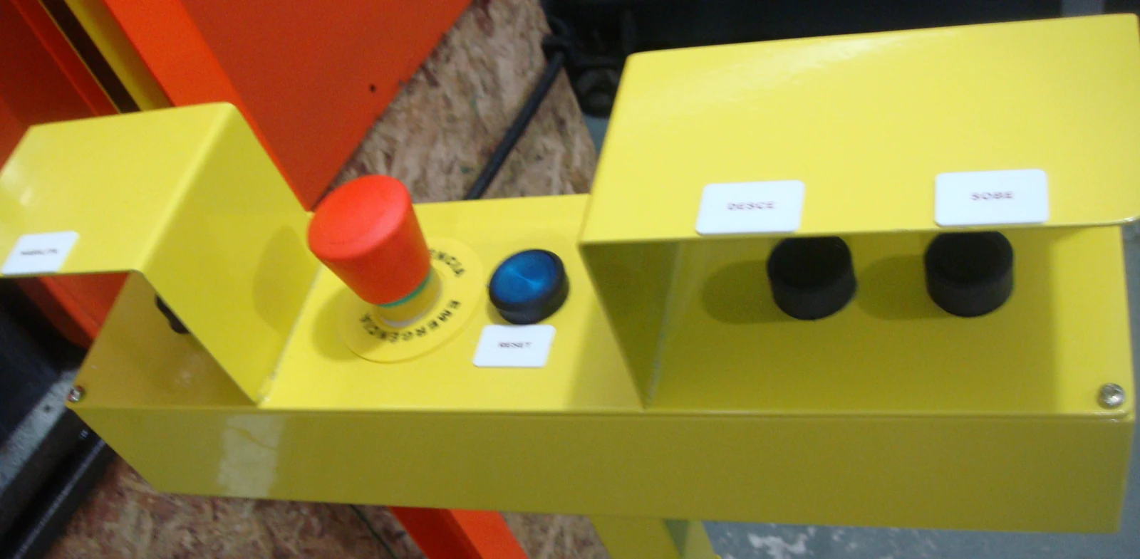 Posto de comando amarelo com dois botões pretos sob anteparos, botão azul e botão vermelho de emergência.